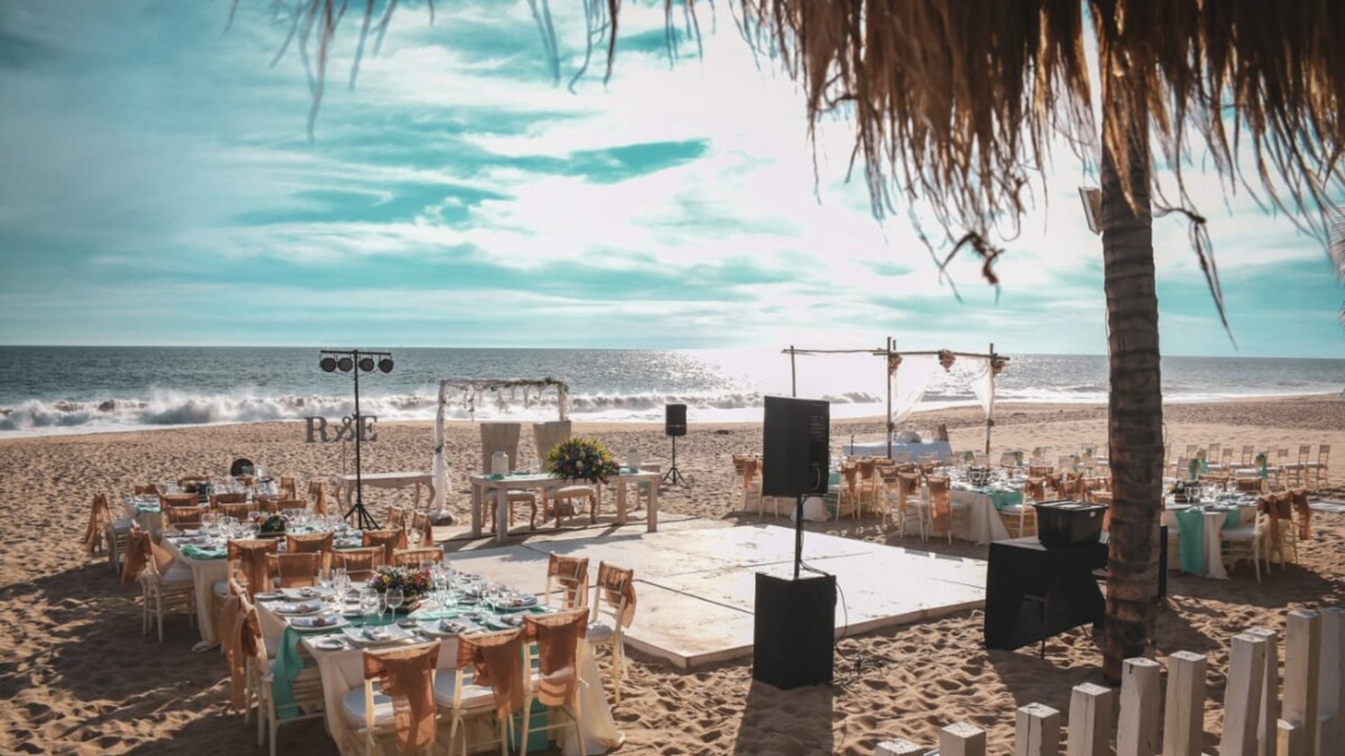 Montaje de boda en la playa con mesas decoradas en menta y dorado, pista de baile y el mar de fondo