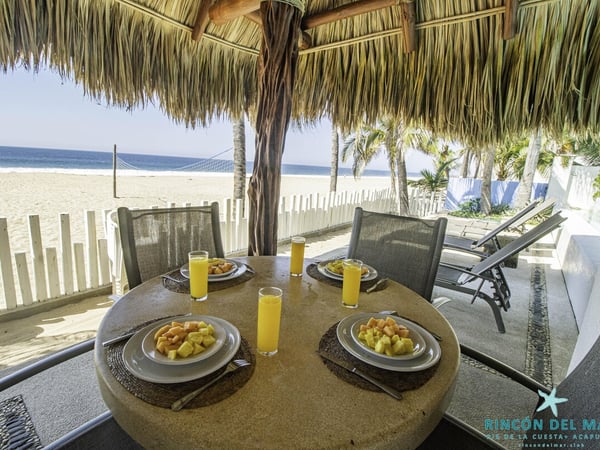 Desayuno tropical bajo palapa frente al mar: mesa con fruta y jugos, playa y océano al fondo
