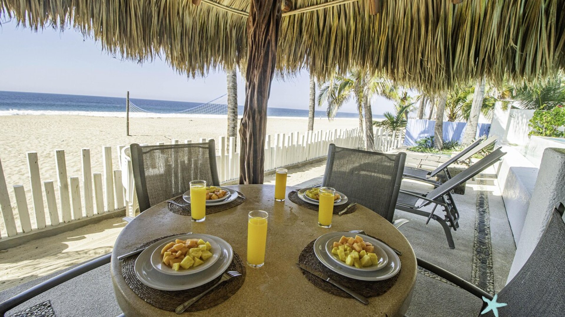 Desayuno tropical bajo palapa frente al mar: mesa con fruta y jugos, playa y océano al fondo