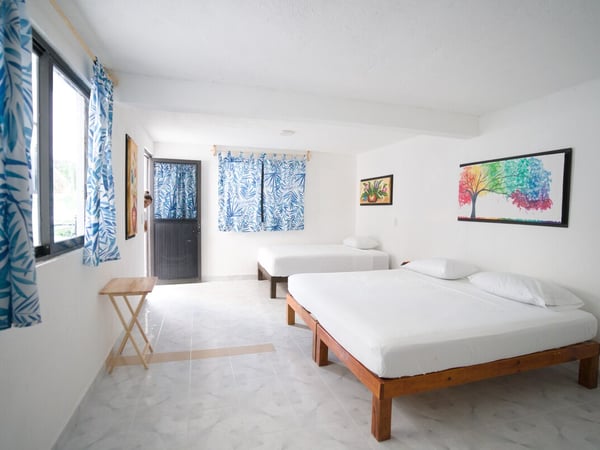 Recámara amplia y luminosa con dos camas individuales de madera, cortinas azules tropicales y arte colorido en paredes blancas