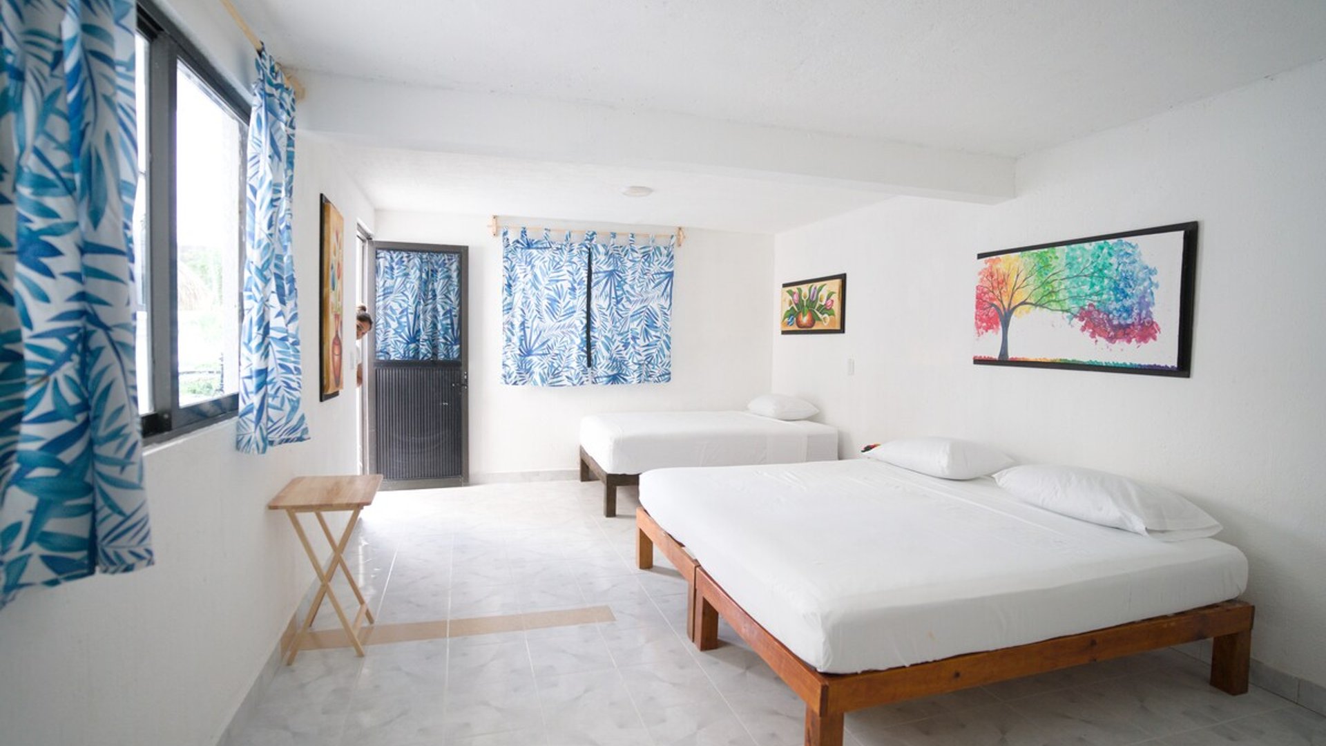Recámara amplia y luminosa con dos camas individuales de madera, cortinas azules tropicales y arte colorido en paredes blancas
