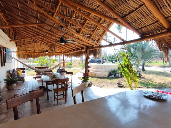 Palapa amplia con techo de palma, comedor exterior de madera, hamaca y vista al jardín con jacuzzi al fondo