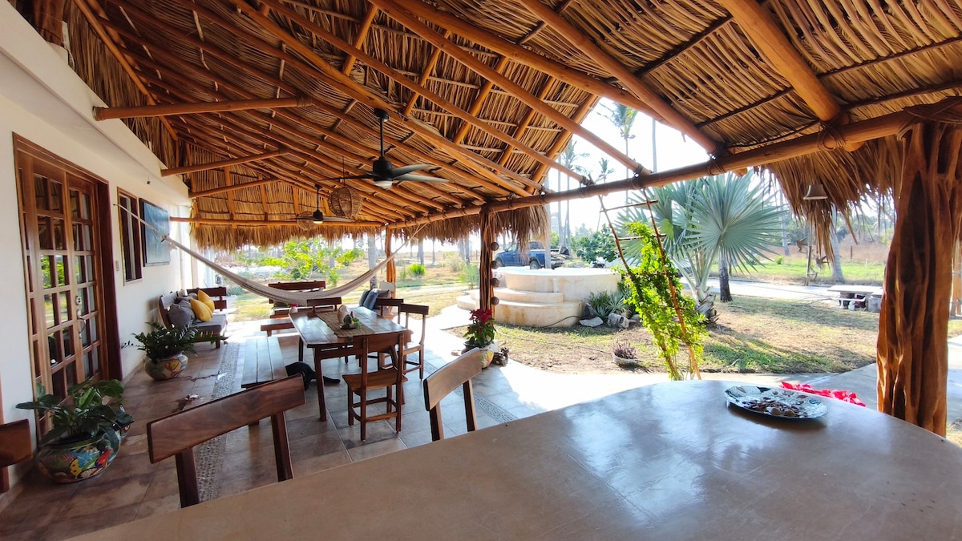Palapa amplia con techo de palma, comedor exterior de madera, hamaca y vista al jardín con jacuzzi al fondo