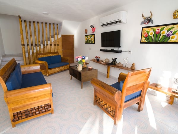 Sala con muebles de madera tallada y cojines azules, decoración artesanal mexicana, escalera de bambú y televisor en pared blanca