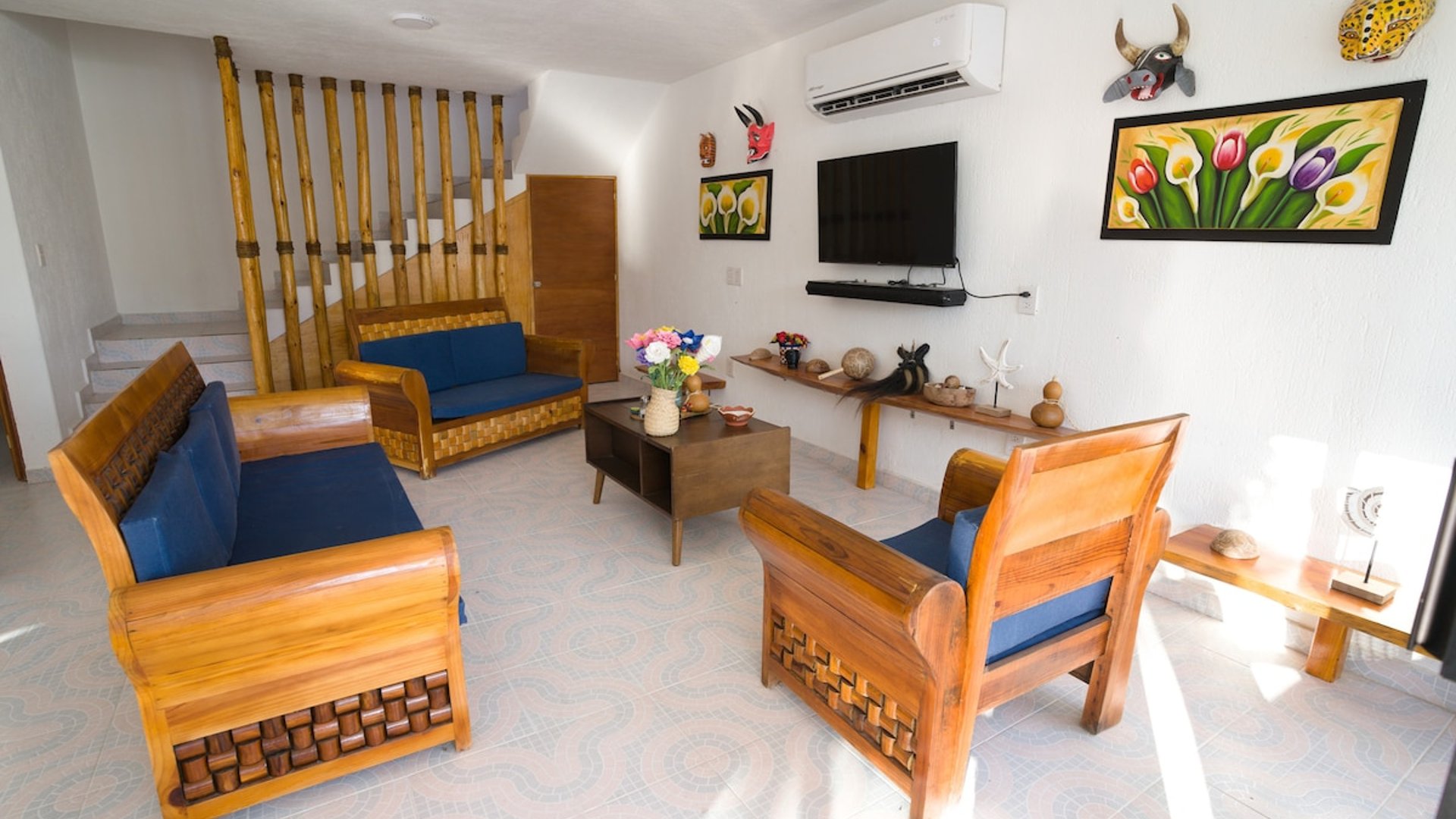 Sala con muebles de madera tallada y cojines azules, decoración artesanal mexicana, escalera de bambú y televisor en pared blanca