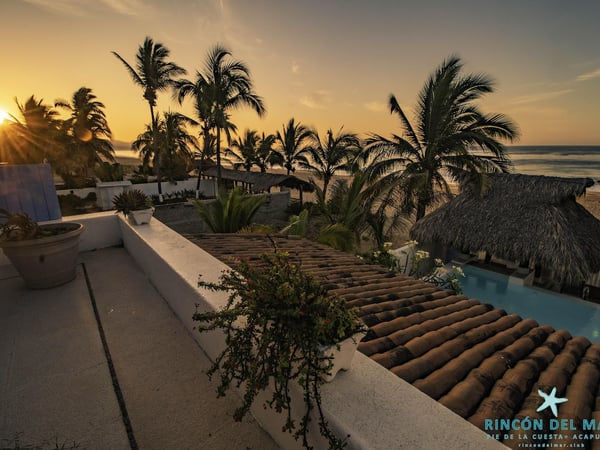 Vista desde terraza con techo de teja al atardecer: alberca, palapa frente al mar y palmeras en silueta