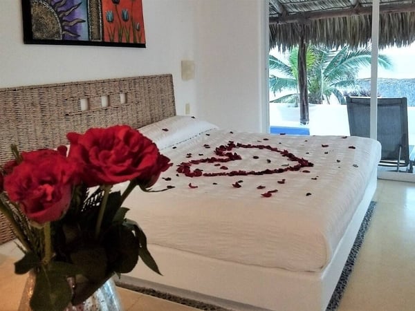 Recámara con cama king decorada con pétalos de rosa en forma de corazón, rosas rojas en primer plano y vista a terraza con palmeras y alberca