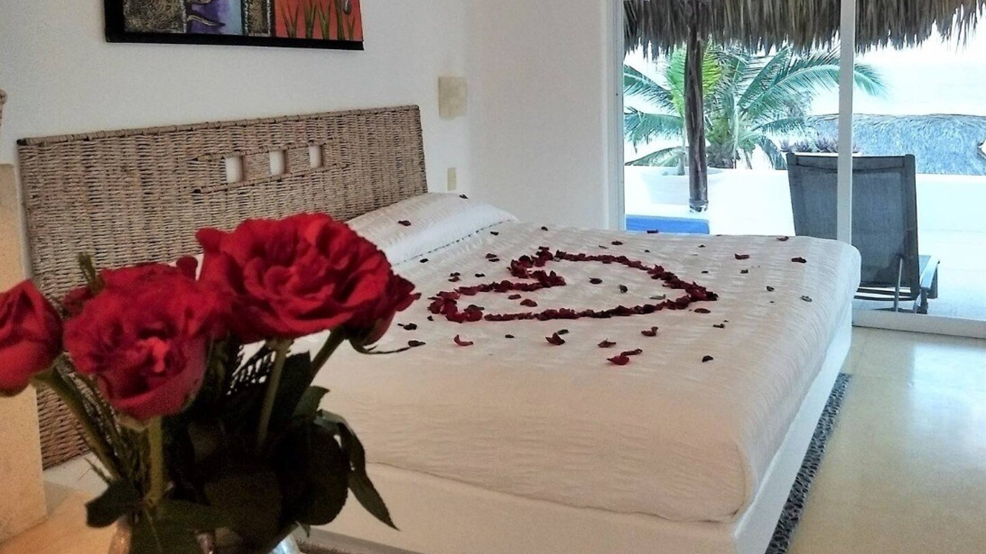 Recámara con cama king decorada con pétalos de rosa en forma de corazón, rosas rojas en primer plano y vista a terraza con palmeras y alberca