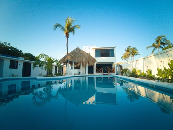 Alberca amplia con reflejo de palapa y villa blanca de dos pisos bajo cielo azul al atardecer