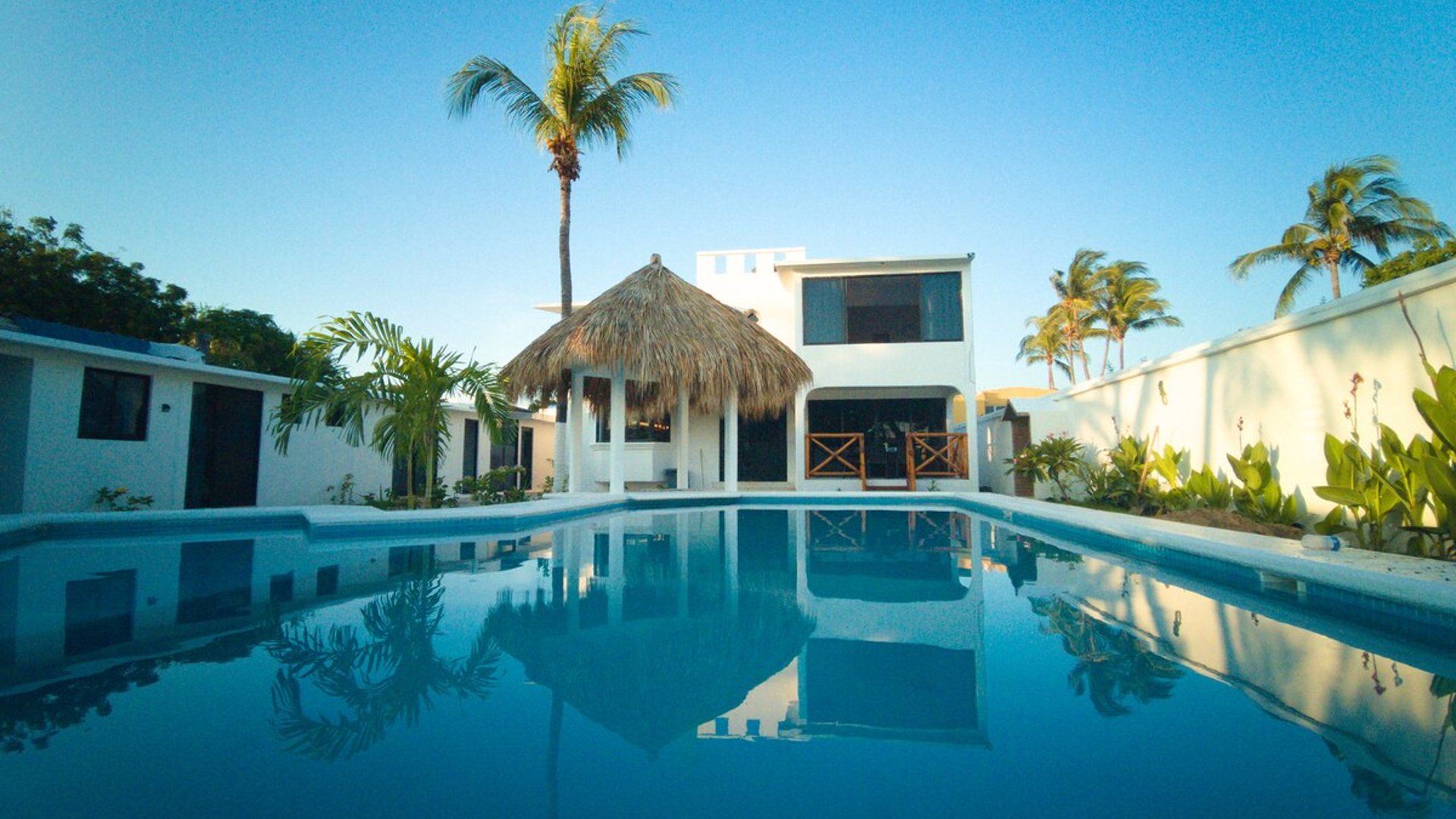 Alberca amplia con reflejo de palapa y villa blanca de dos pisos bajo cielo azul al atardecer