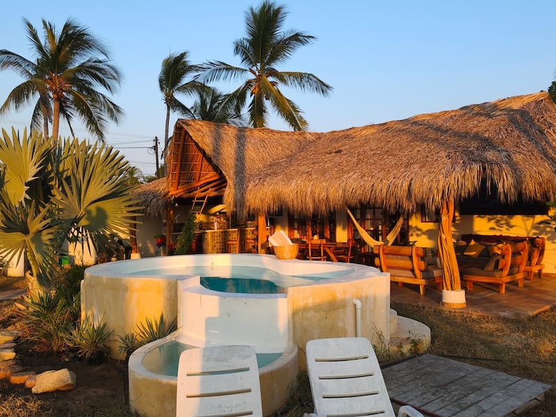 Jacuzzi circular y palapa rústica con hamaca al atardecer, rodeados de palmeras tropicales