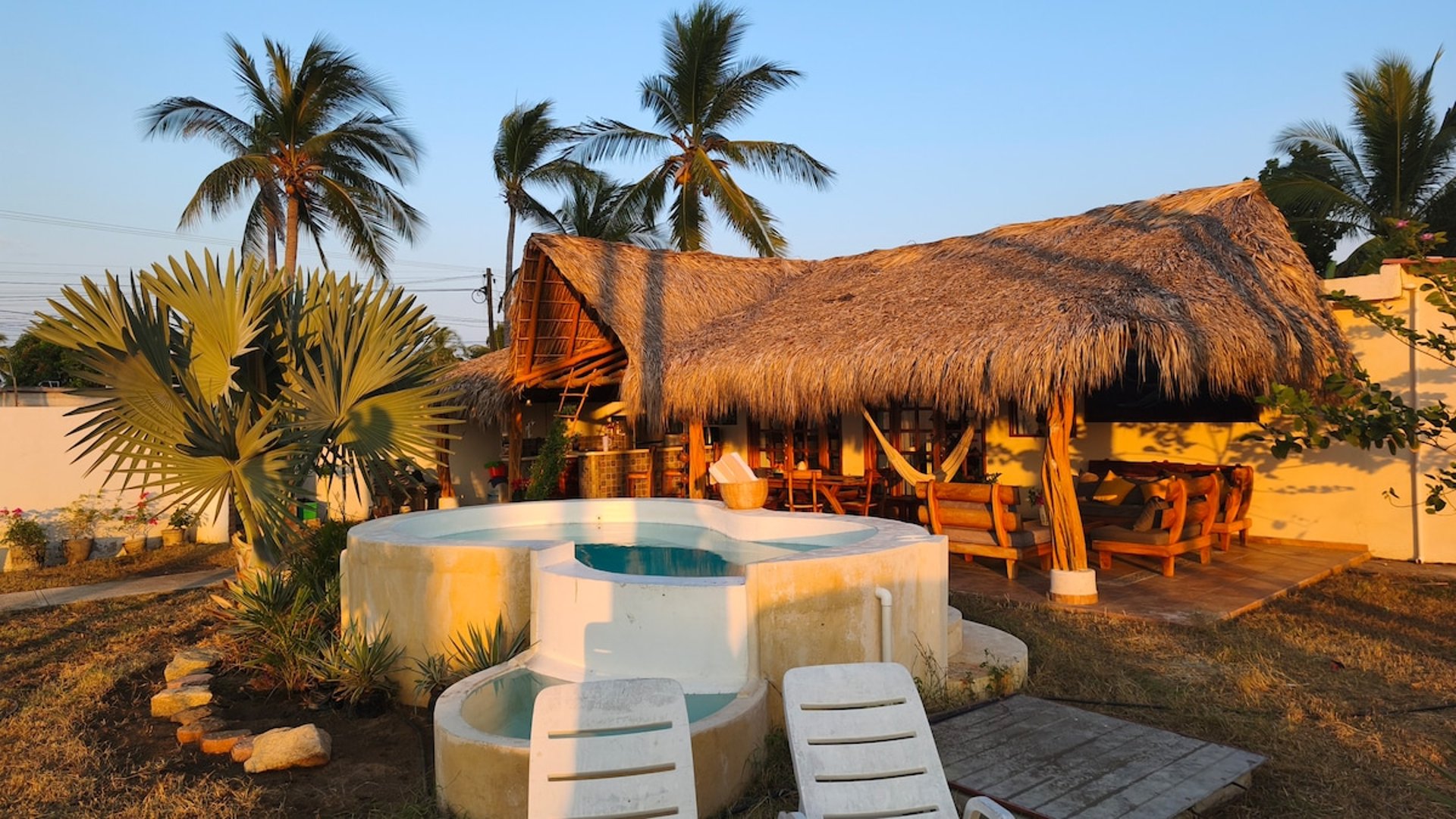 Jacuzzi circular y palapa rústica con hamaca al atardecer, rodeados de palmeras tropicales