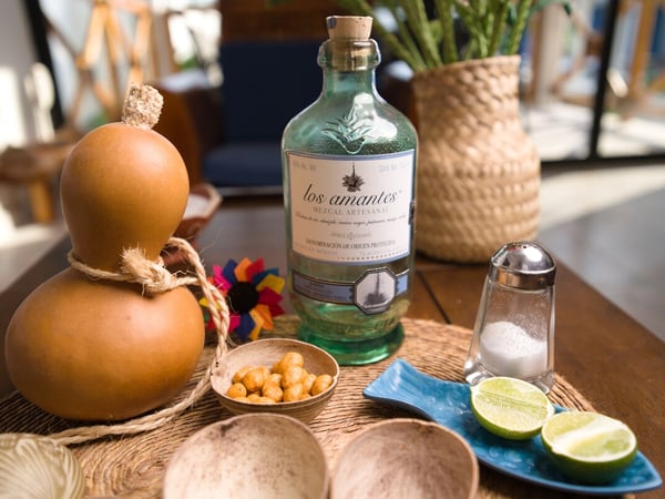 Botella de mezcal artesanal Los Amantes con jícara, limones, sal y botanas sobre mesa de madera con ambiente cálido