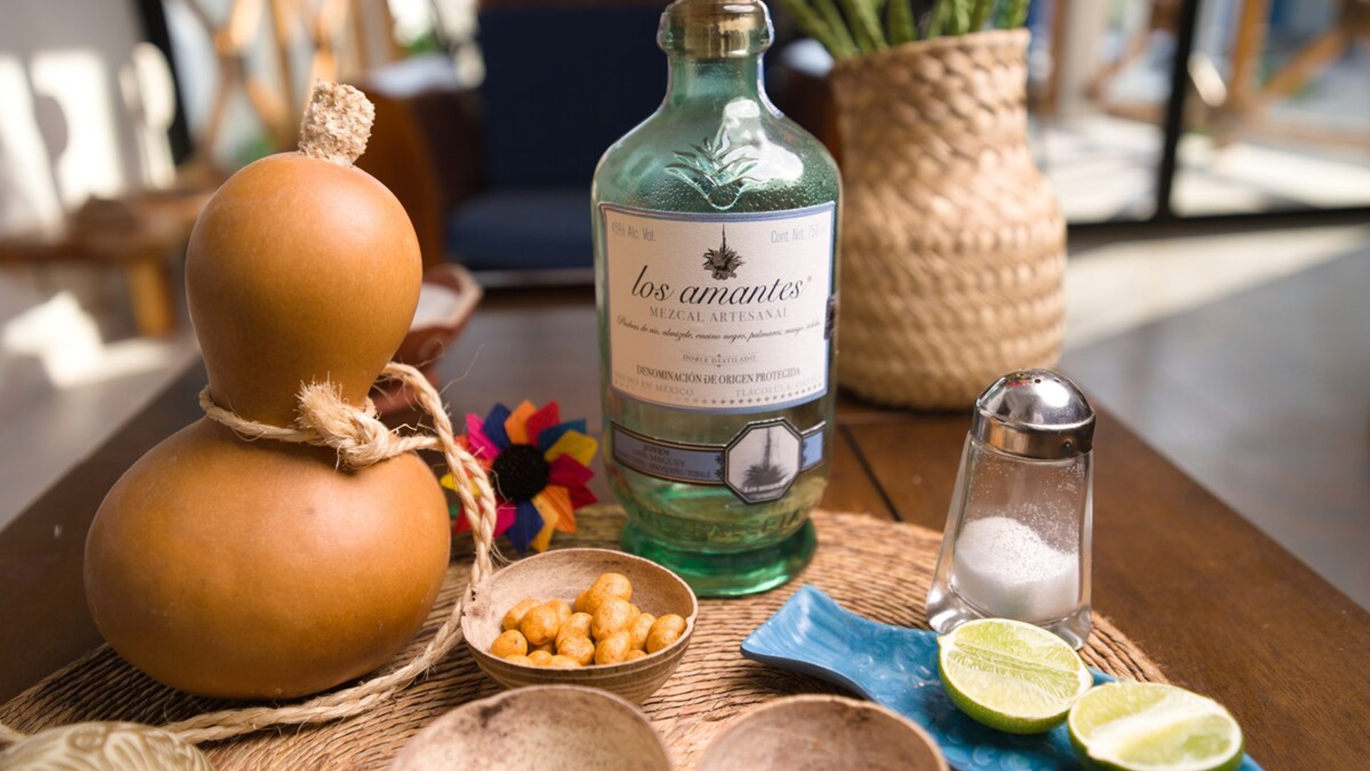 Botella de mezcal artesanal Los Amantes con jícara, limones, sal y botanas sobre mesa de madera con ambiente cálido