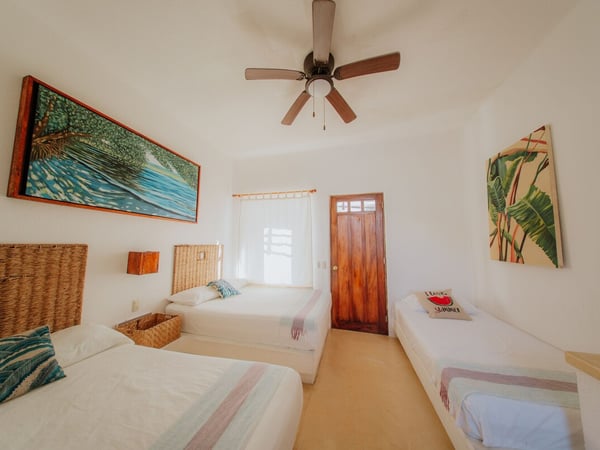Recámara triple con tres camas, cabeceras de mimbre, arte tropical en paredes blancas y ventilador de techo