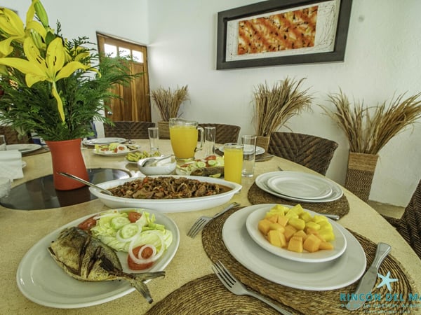 Mesa del comedor servida con pescado, frutas, guisos y jugo de naranja en ambiente interior luminoso
