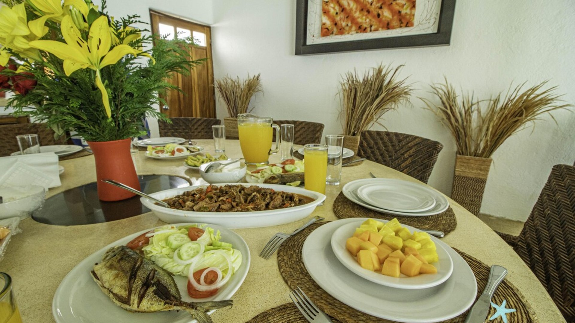 Mesa del comedor servida con pescado, frutas, guisos y jugo de naranja en ambiente interior luminoso