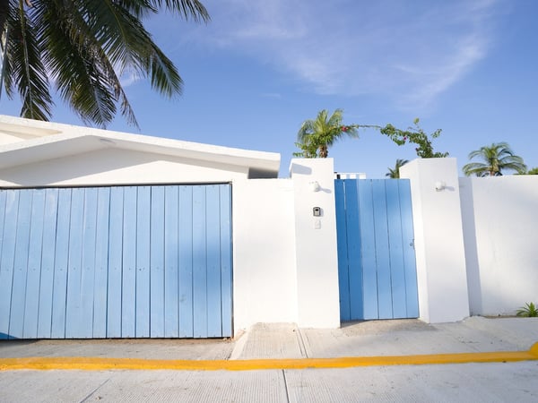 Fachada blanca con portón y puerta peatonal en azul cielo, palmeras asomando bajo cielo despejado