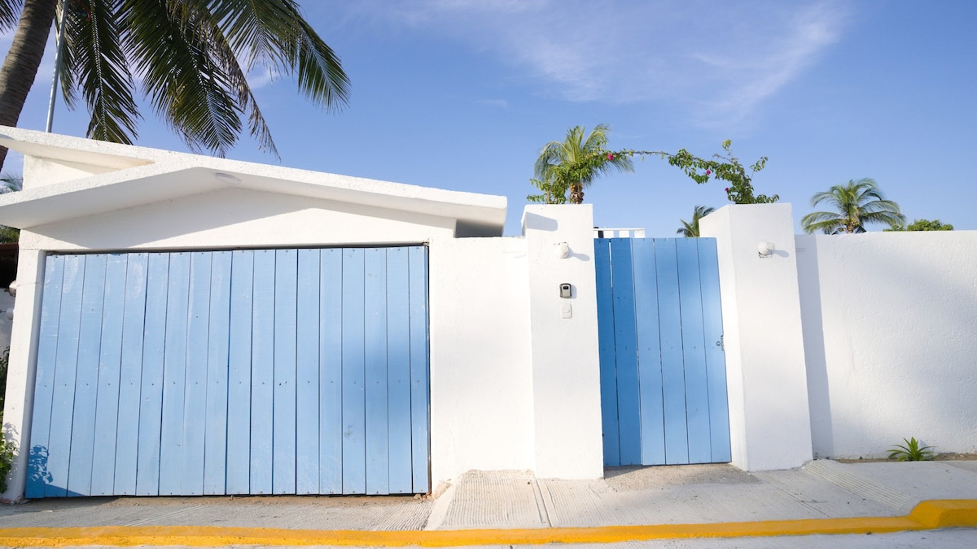 Fachada blanca con portón y puerta peatonal en azul cielo, palmeras asomando bajo cielo despejado