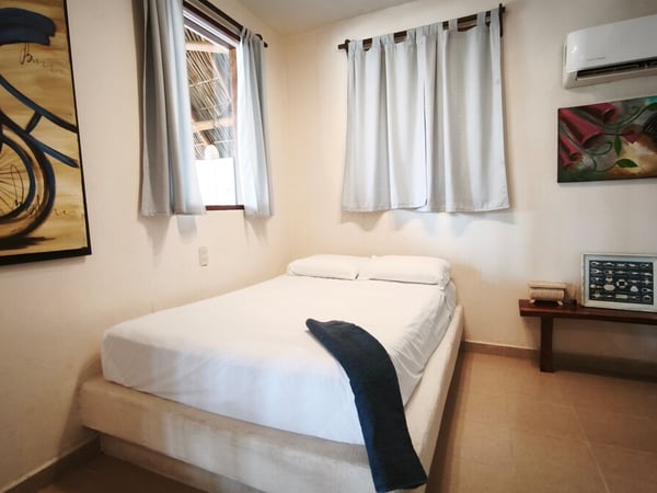 Recámara sencilla con cama doble en ropa blanca, pinturas coloridas en paredes y aire acondicionado mini-split