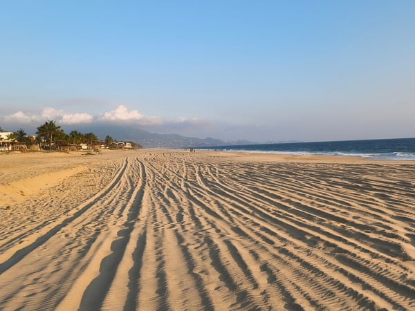 Amplia playa de arena dorada con huellas onduladas al amanecer, mar azul y montañas al fondo