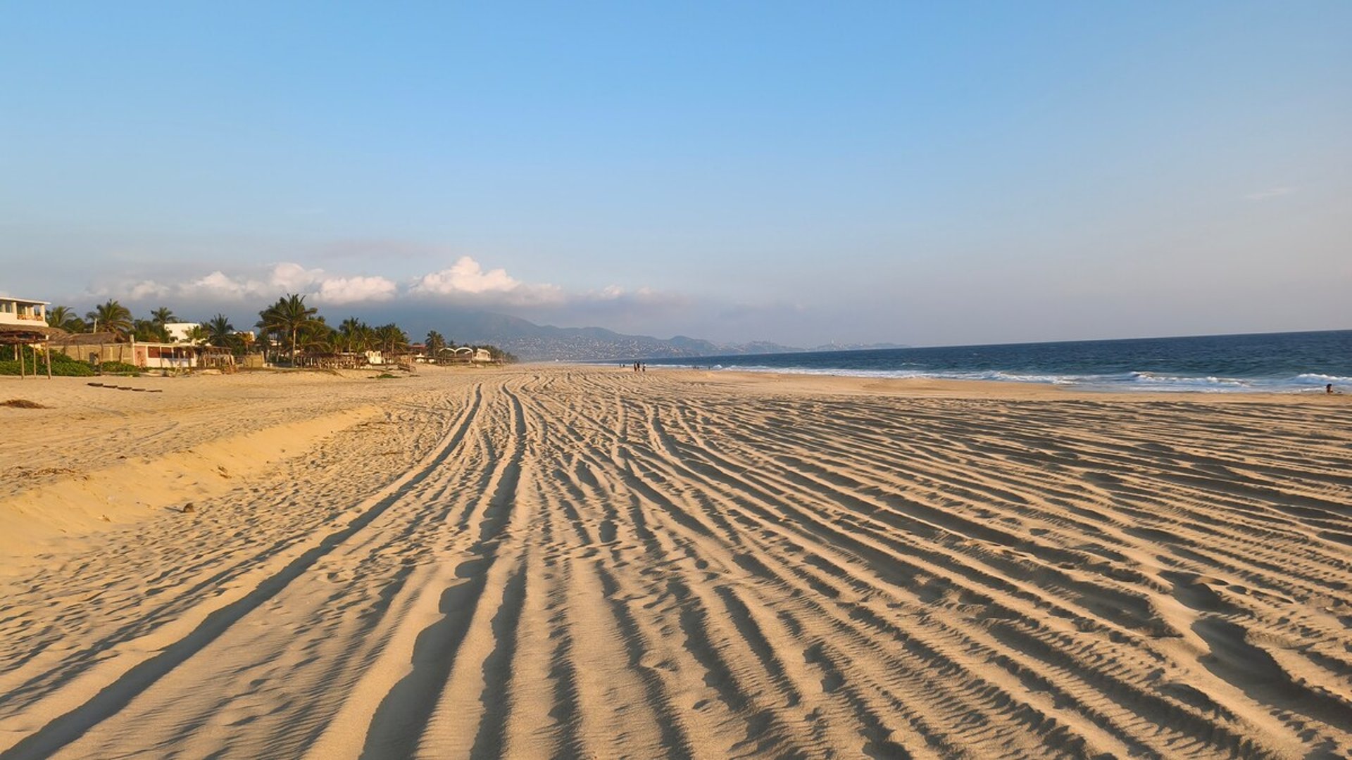 Amplia playa de arena dorada con huellas onduladas al amanecer, mar azul y montañas al fondo