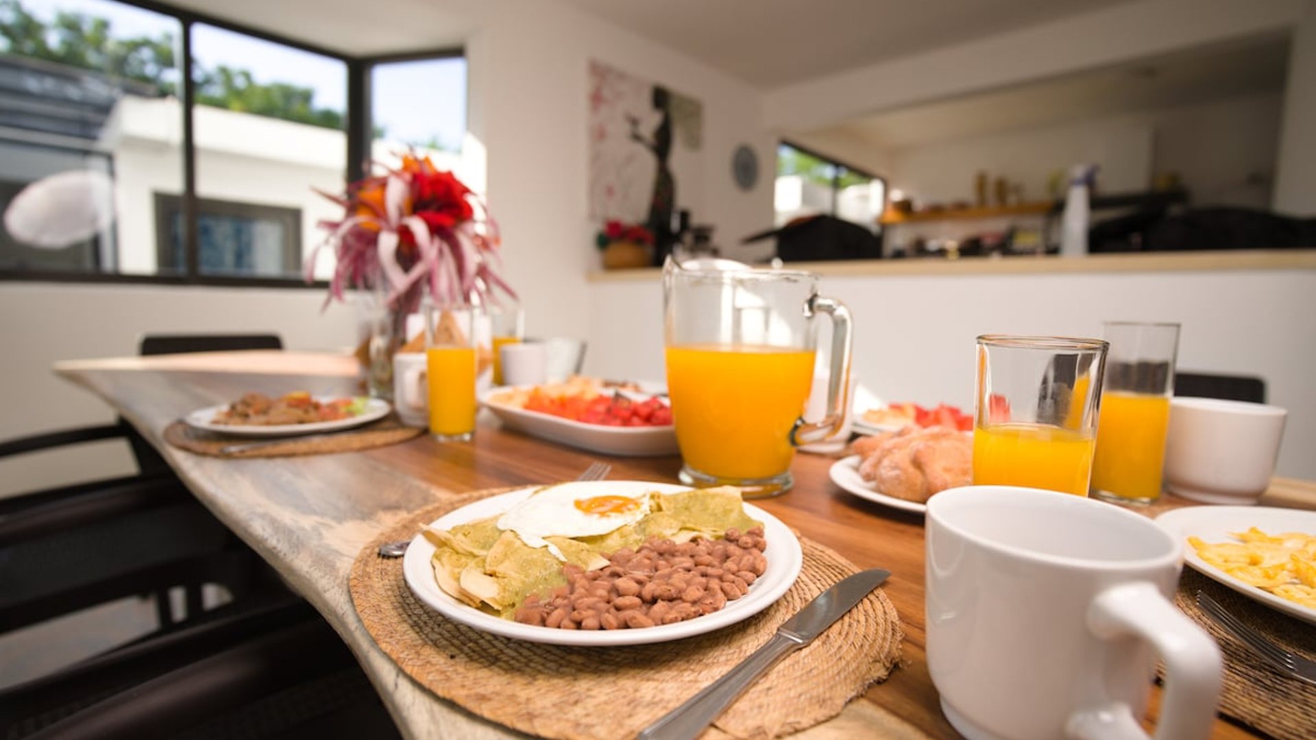 Mesa de madera con desayuno mexicano servido: chilaquiles, frijoles, fruta y jugo de naranja en comedor luminoso con cocina al fondo