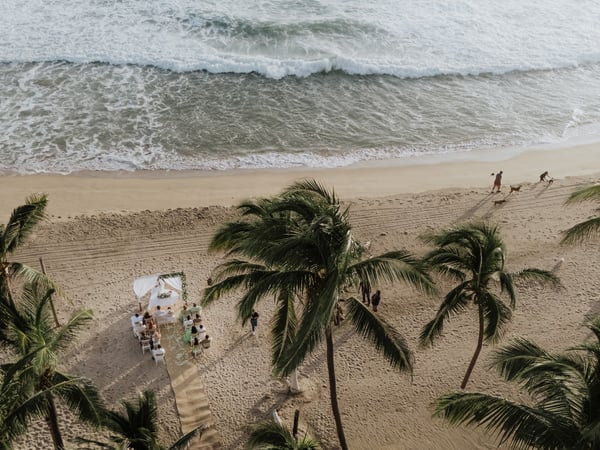 Vista aérea de ceremonia de boda en playa con sillas y arco blanco entre palmeras frente al mar