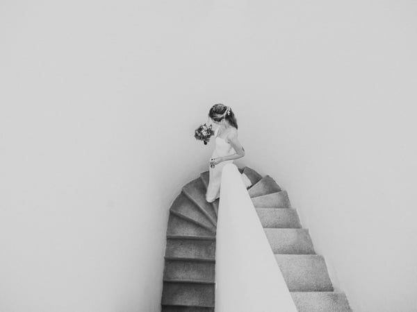 Novia descendiendo escalera curva de villa con vestido y ramo en blanco y negro