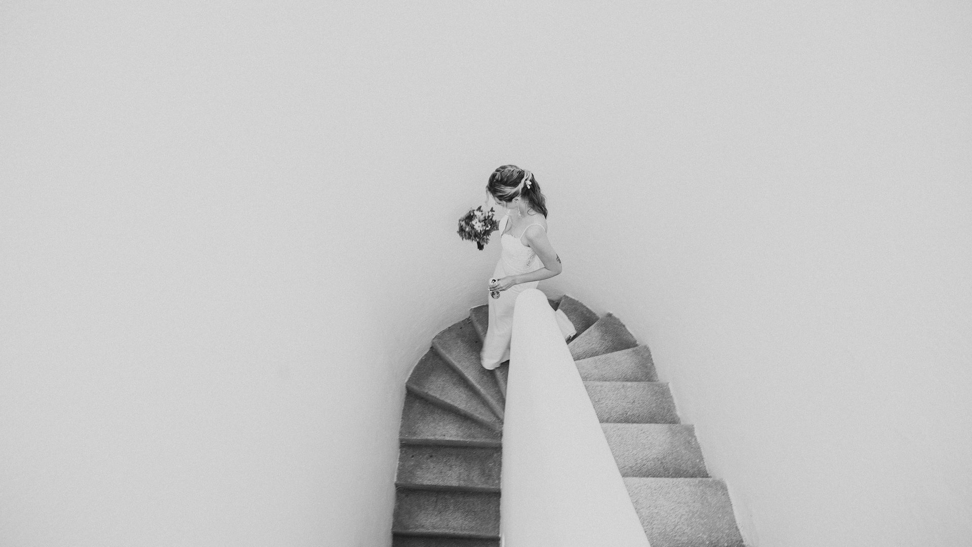 Novia descendiendo escalera curva de villa con vestido y ramo en blanco y negro