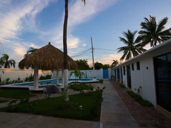 Jardín al atardecer con palapa, alberca y andador junto a habitaciones de fachada blanca moderna
