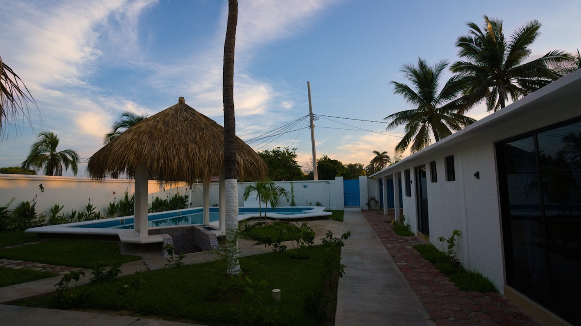 Jardín al atardecer con palapa, alberca y andador junto a habitaciones de fachada blanca moderna