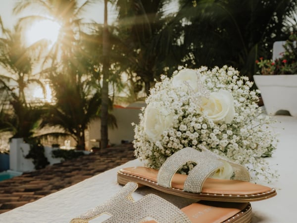 Detalle de sandalias de novia junto a ramo blanco con palmeras tropicales de fondo al atardecer