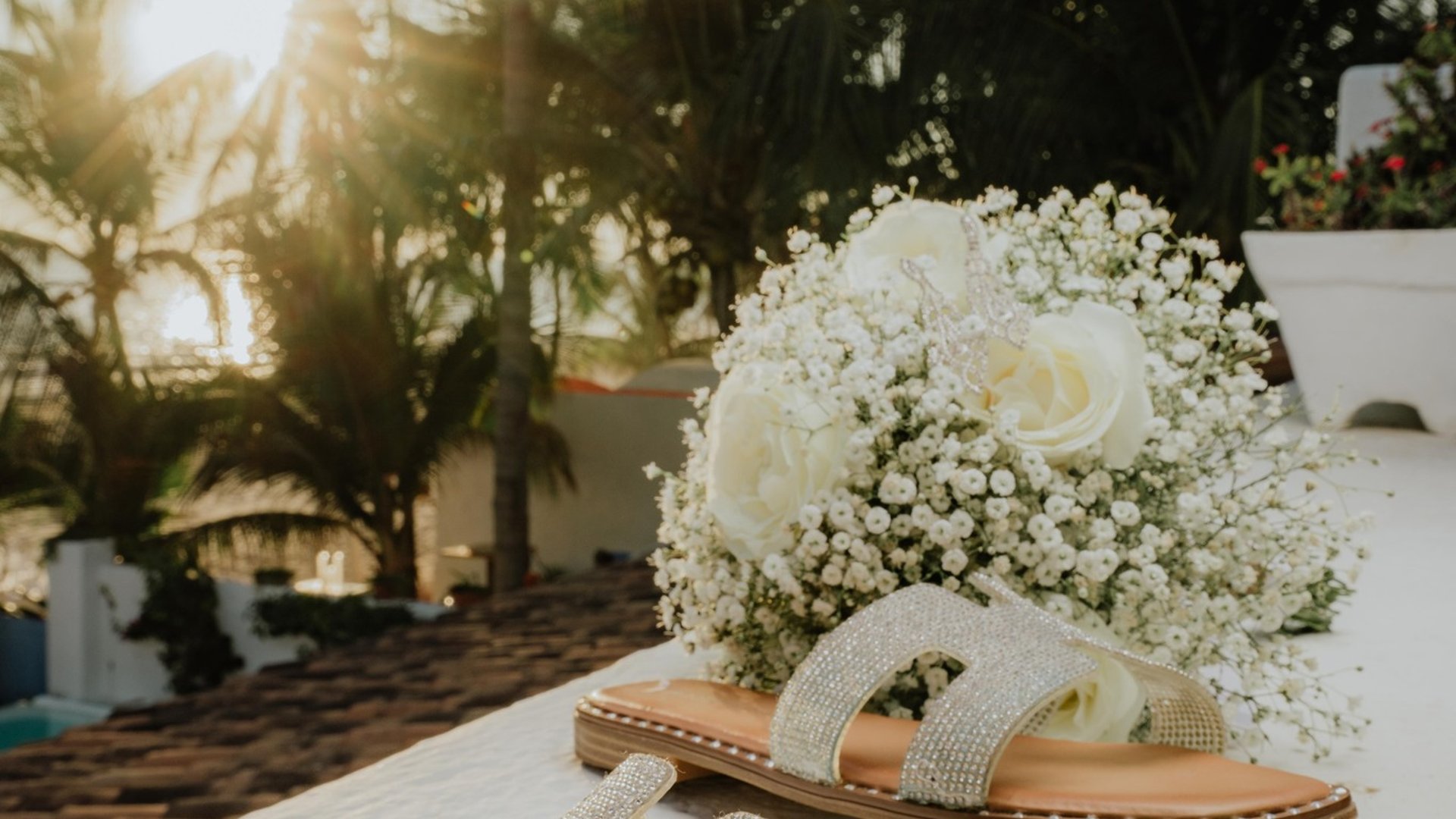 Detalle de sandalias de novia junto a ramo blanco con palmeras tropicales de fondo al atardecer