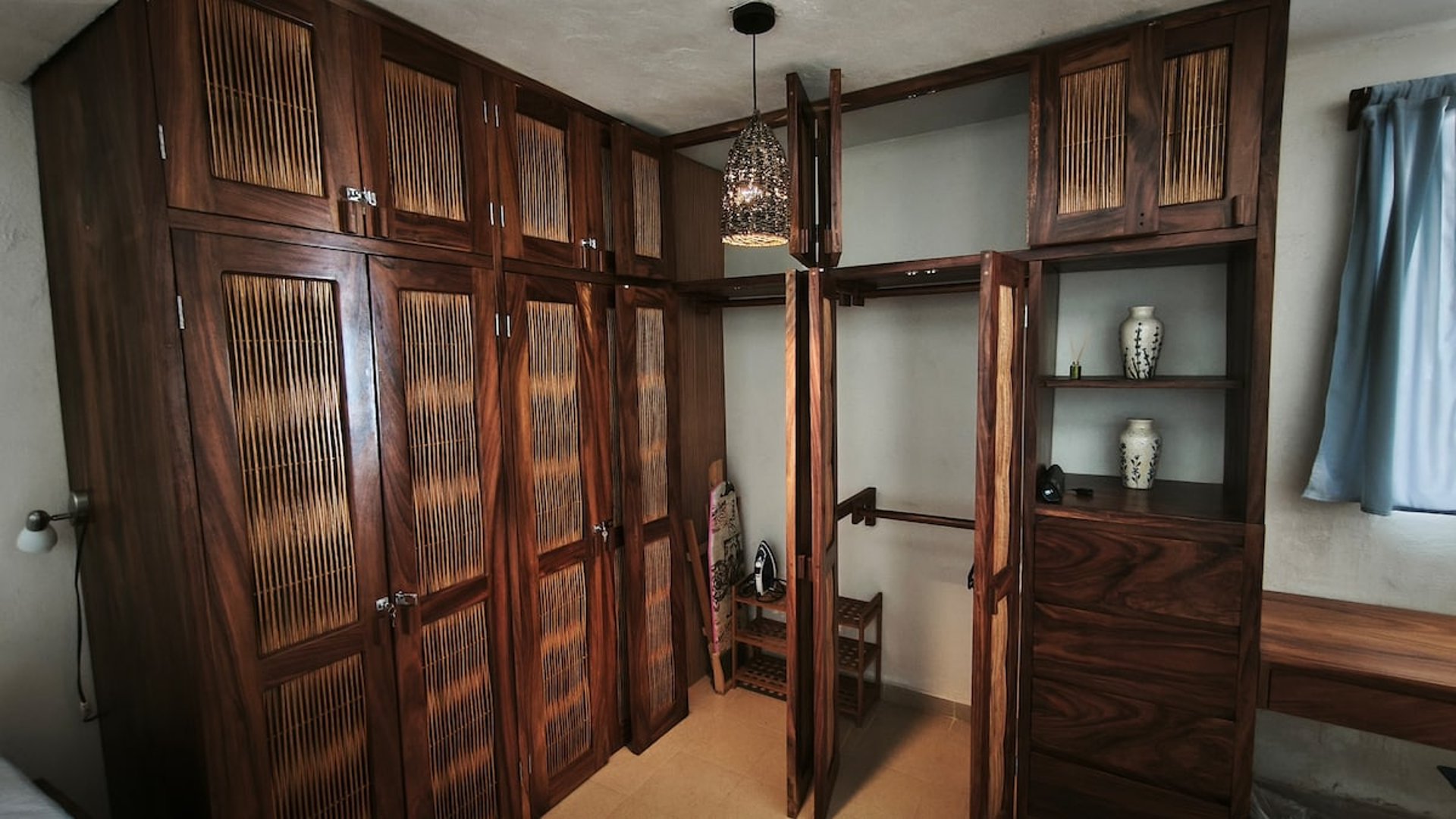 Amplio vestidor con armarios de madera oscura y paneles de bambú, lámpara artesanal y repisas decorativas