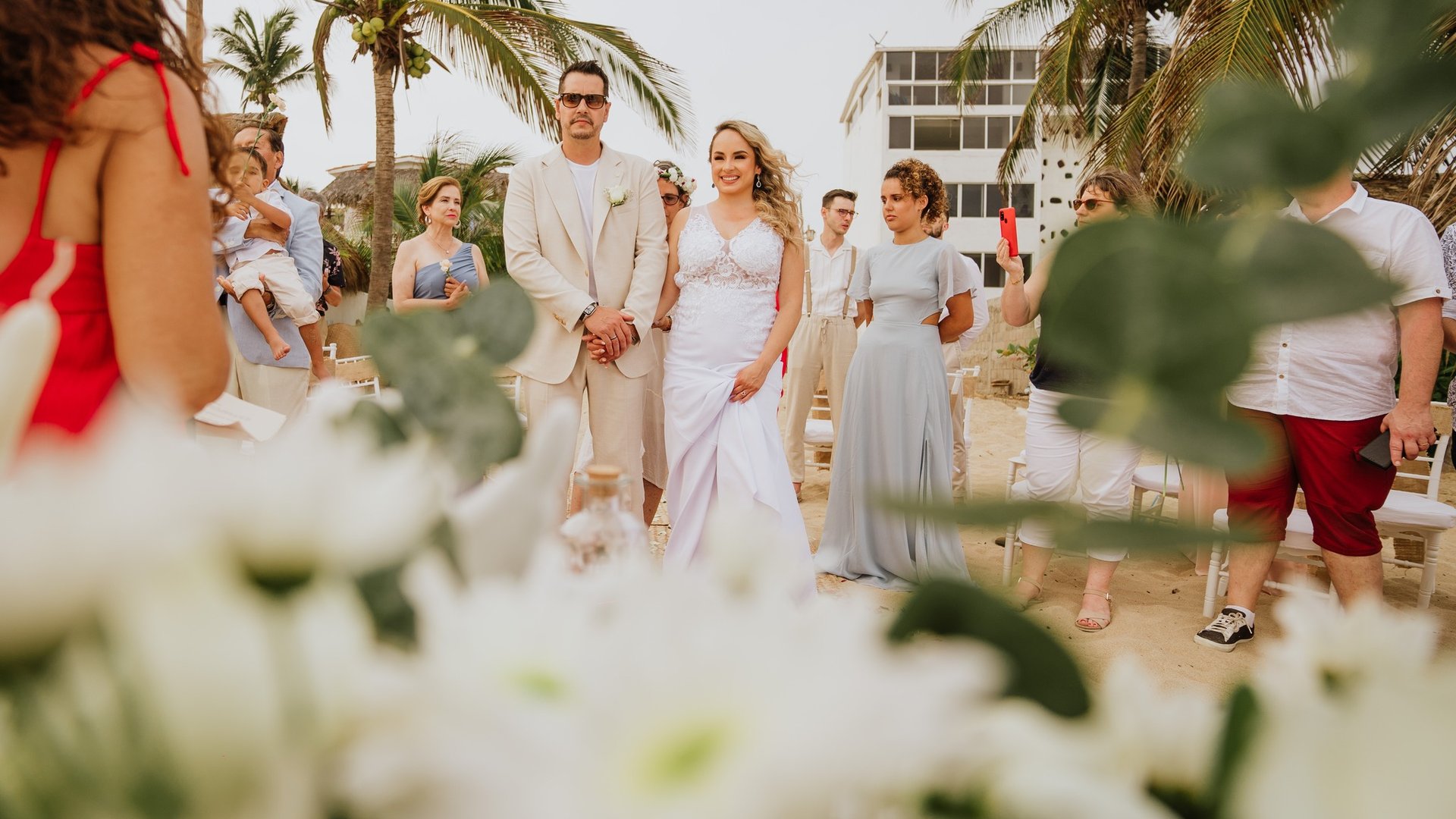 Ceremonia de boda en playa con flores blancas, palmeras tropicales e invitados en arena