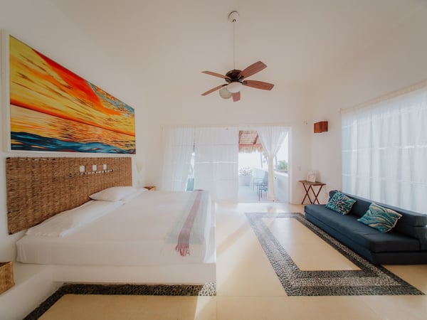 Recámara amplia y luminosa con cama king, sofá azul, piso de mosaico y acceso a terraza con palapa al fondo