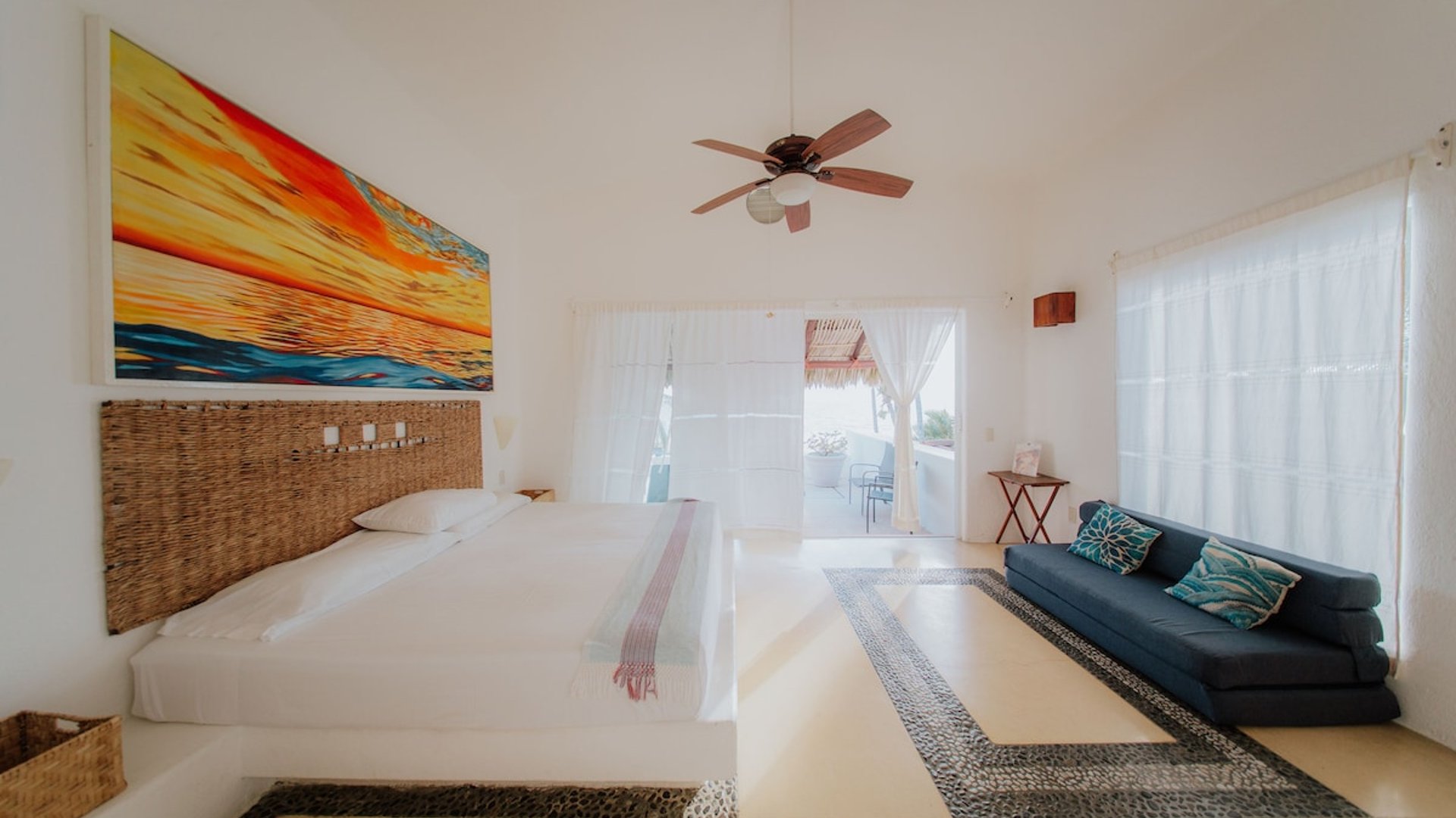 Recámara amplia y luminosa con cama king, sofá azul, piso de mosaico y acceso a terraza con palapa al fondo