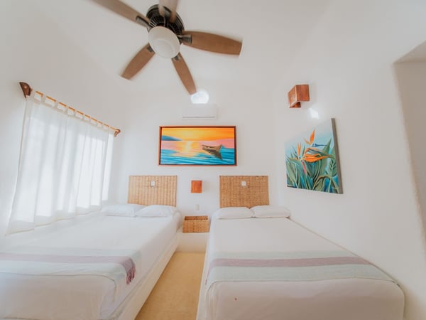 Recámara doble con dos camas individuales, cabeceras de mimbre, pinturas tropicales y ventilador de techo