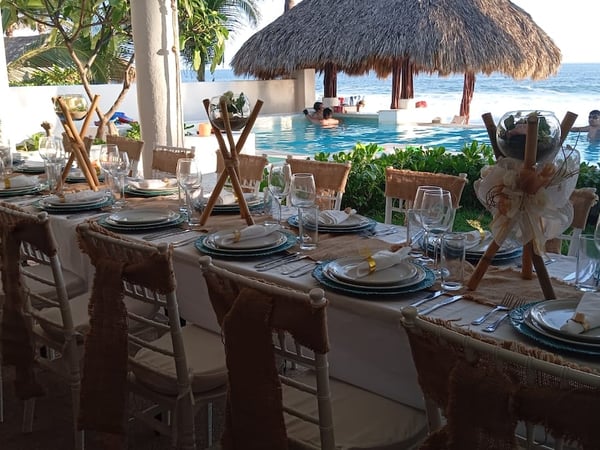 Mesa de banquete decorada con yute y cristalería frente a alberca infinity, palapa y mar al fondo