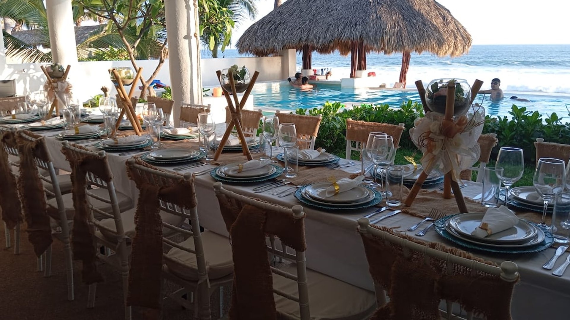 Mesa de banquete decorada con yute y cristalería frente a alberca infinity, palapa y mar al fondo