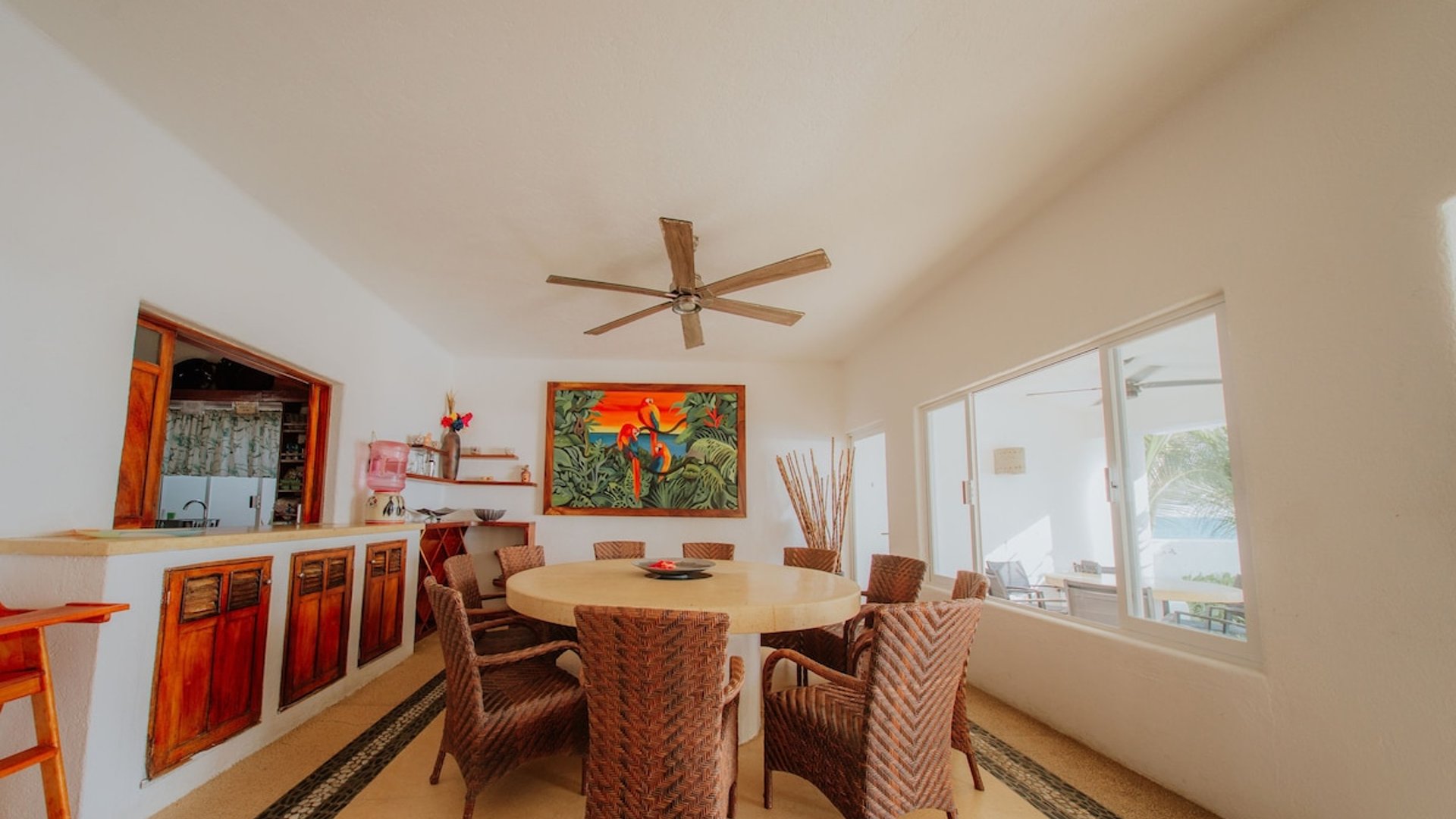 Comedor interior con mesa redonda y sillas de mimbre, barra de cocina y cuadro tropical colorido en pared blanca