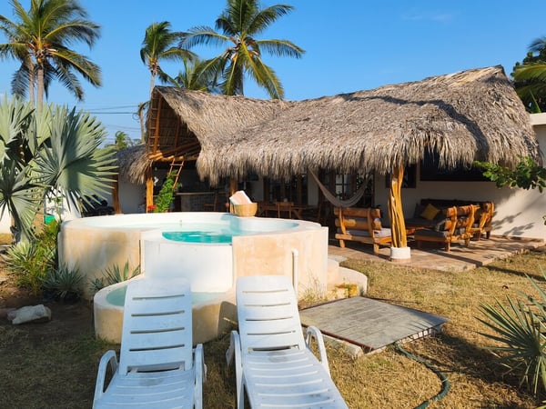 Jardín con jacuzzi redondo, camastros blancos y palapa de palma rodeada de palmeras en luz dorada