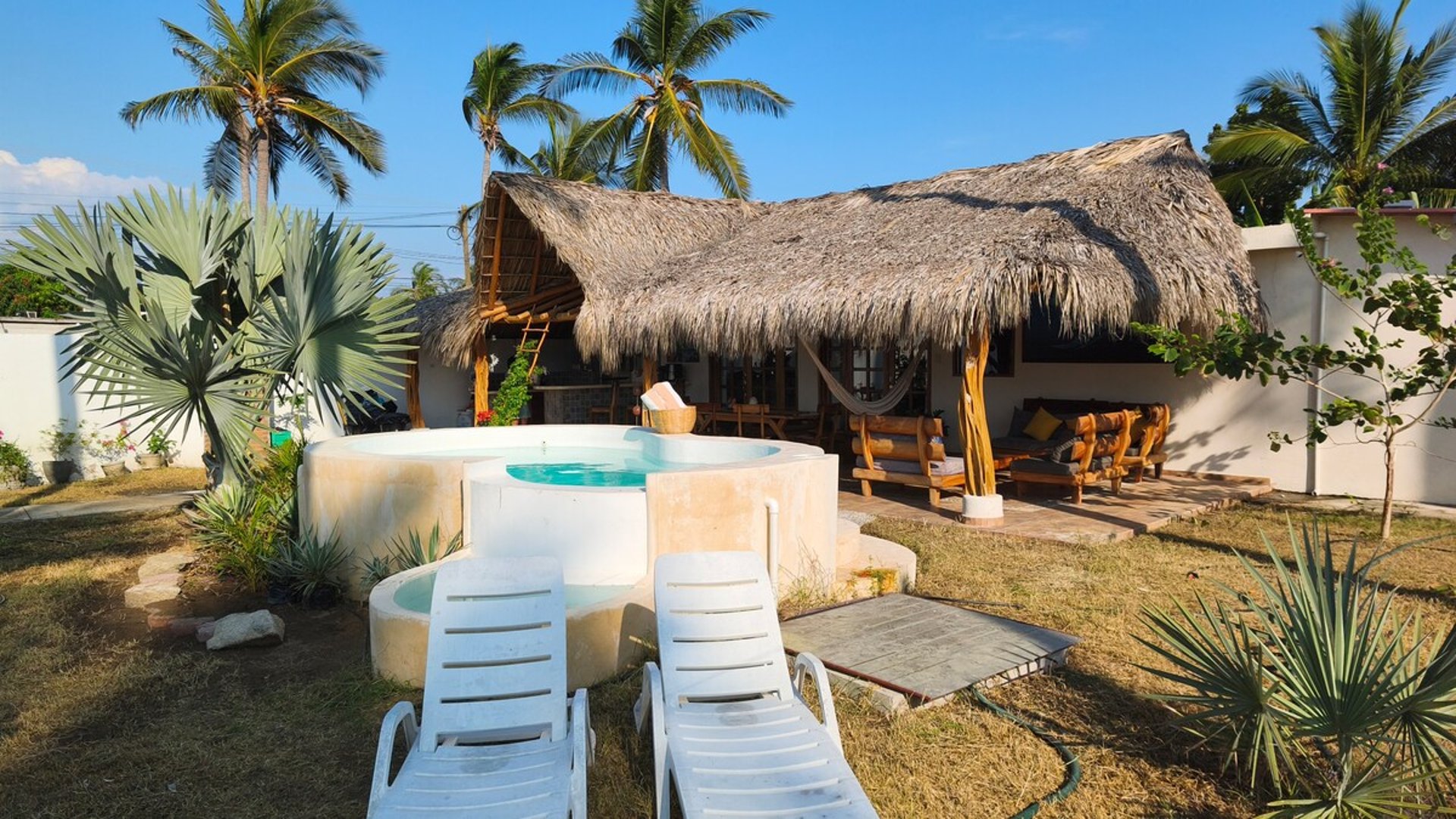 Jardín con jacuzzi redondo, camastros blancos y palapa de palma rodeada de palmeras en luz dorada