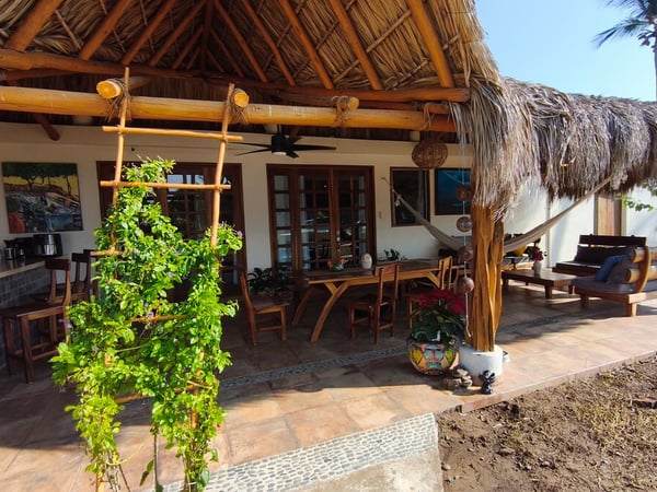 Terraza bajo palapa con techo de palma, comedor exterior de madera, hamaca y sala lounge rodeados de vegetación tropical en luz de día