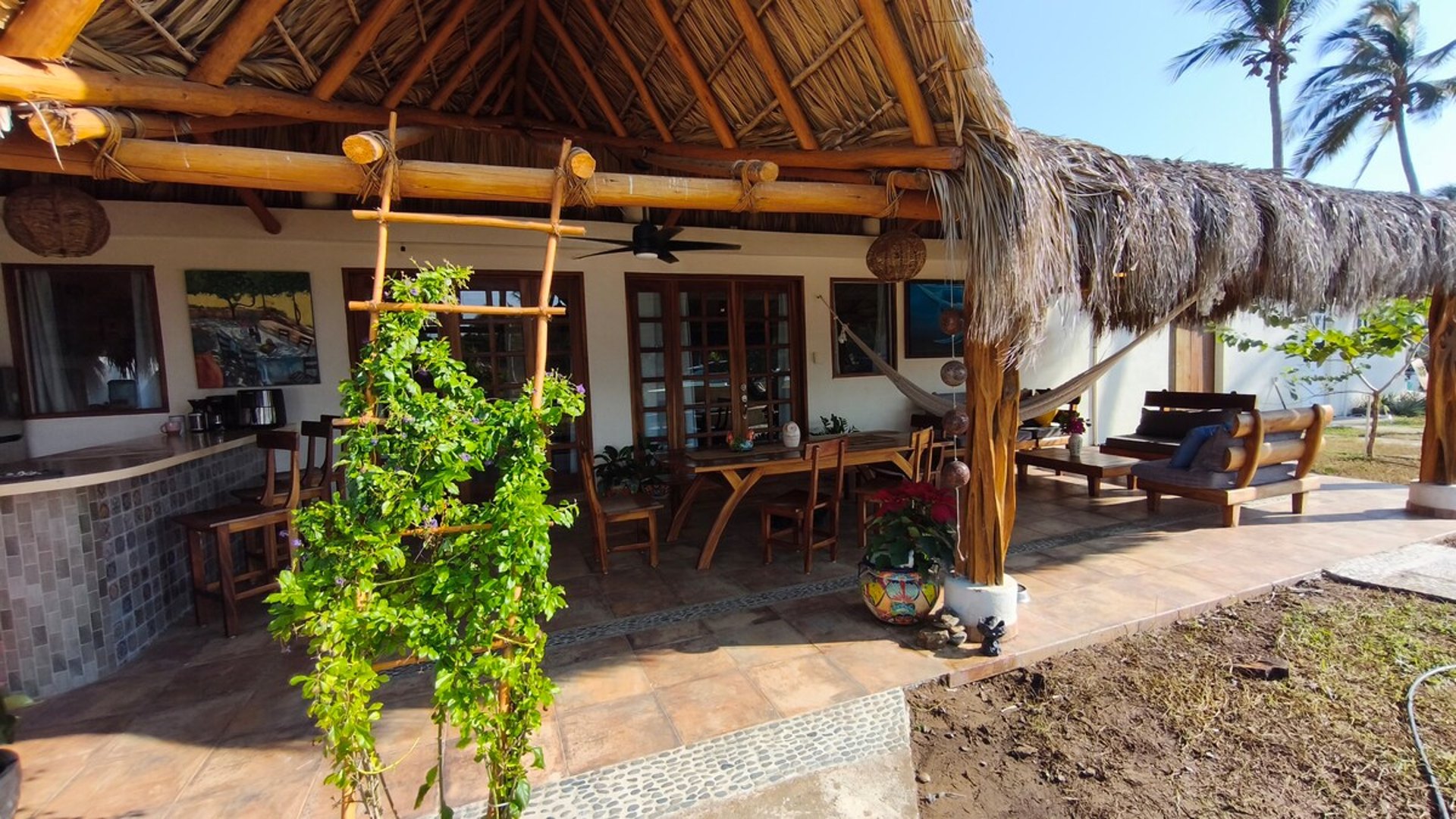 Terraza bajo palapa con techo de palma, comedor exterior de madera, hamaca y sala lounge rodeados de vegetación tropical en luz de día