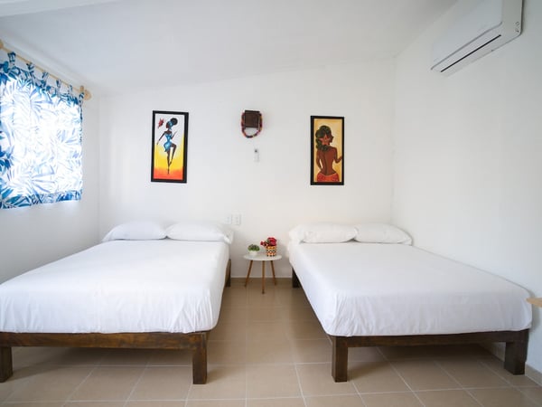 Recámara con dos camas individuales, ropa blanca, cuadros tropicales y aire acondicionado en habitación luminosa