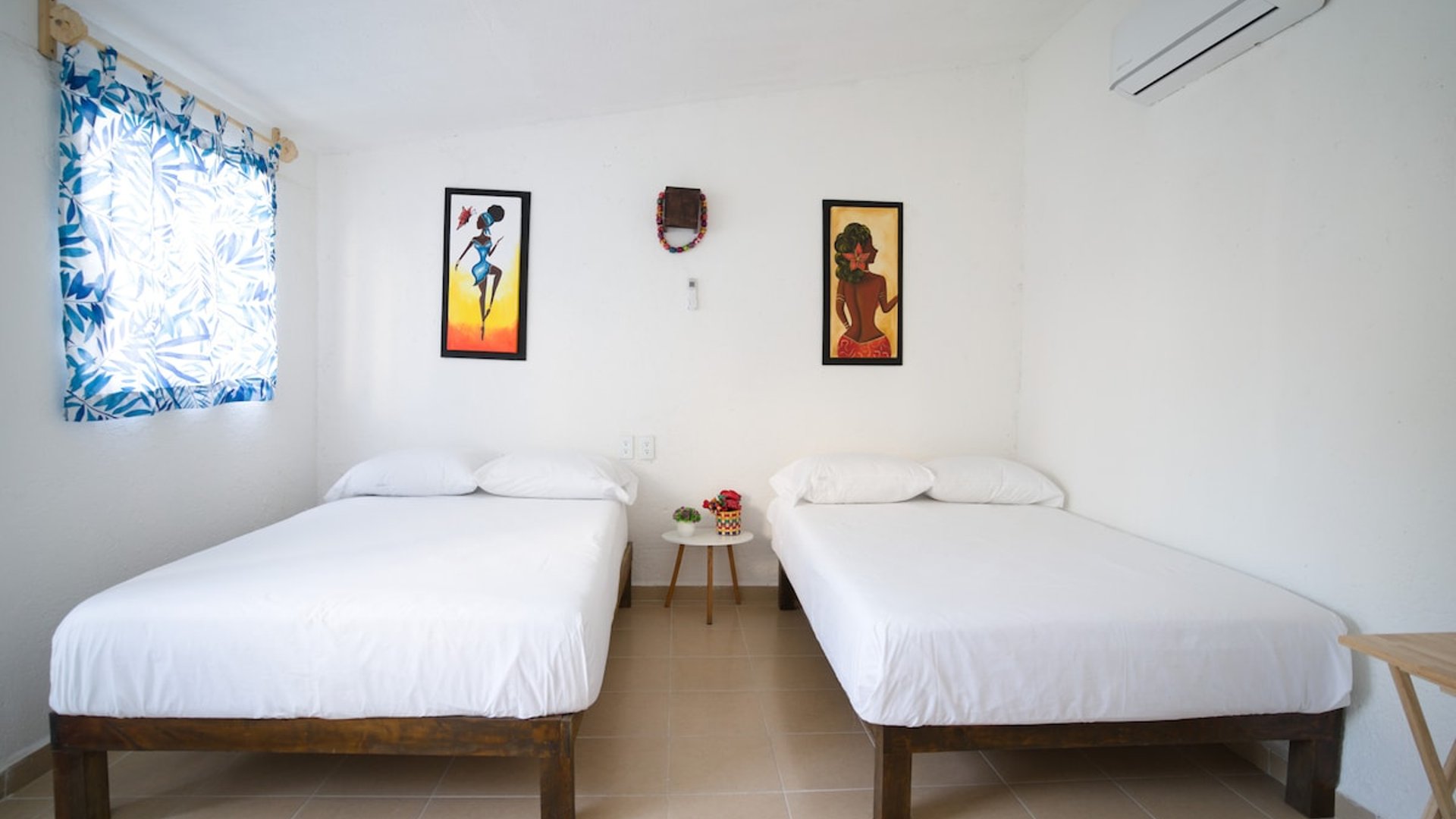Recámara con dos camas individuales, ropa blanca, cuadros tropicales y aire acondicionado en habitación luminosa