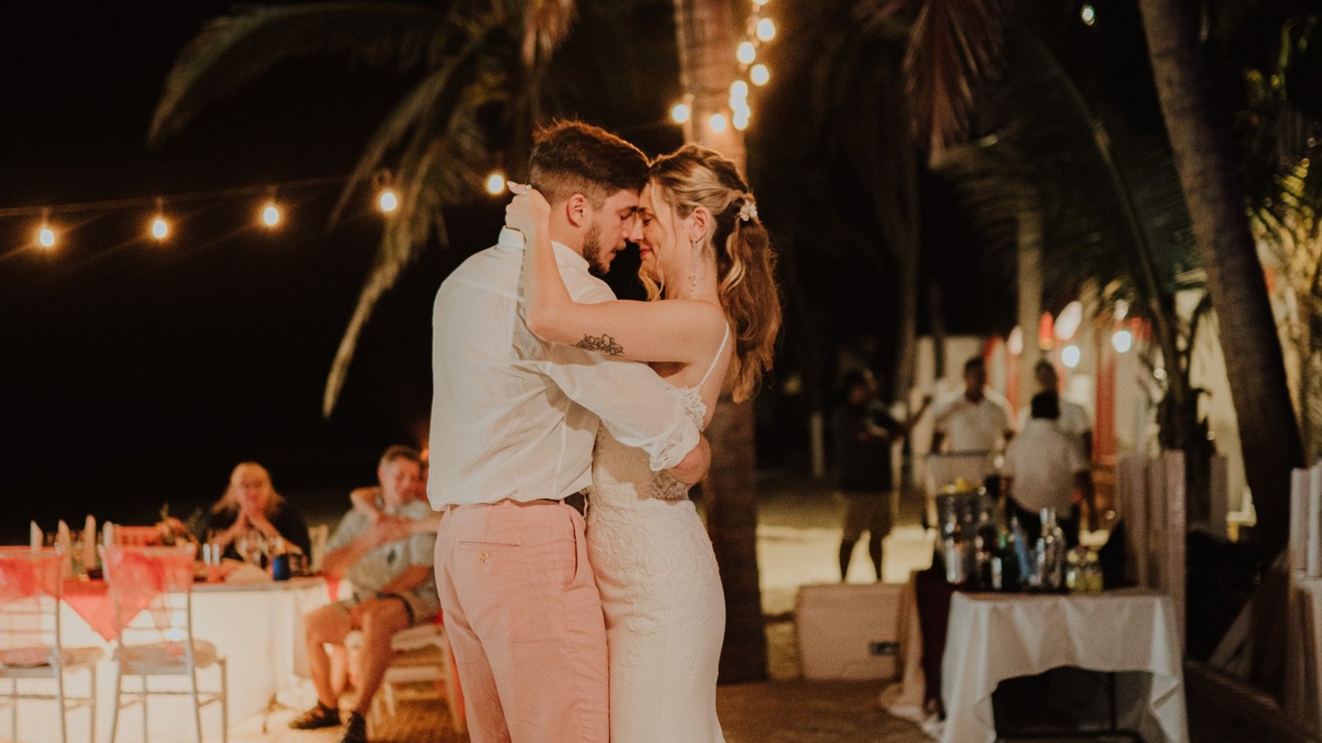 Primer baile de boda en pista bajo palmeras con luces colgantes y bar al fondo