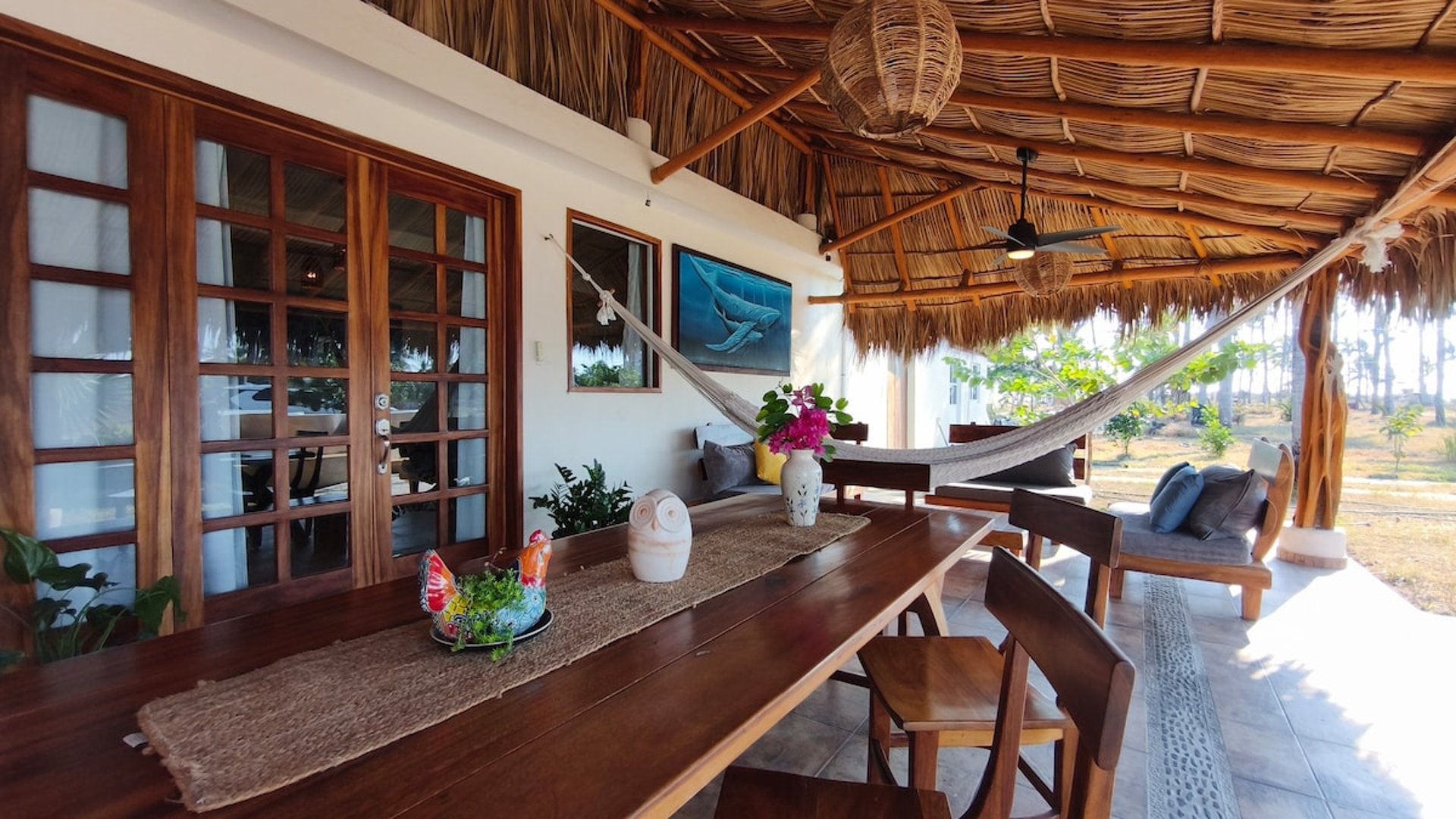 Terraza con palapa, mesa de madera, hamaca y decoración artesanal con vista al jardín y palmeras al fondo
